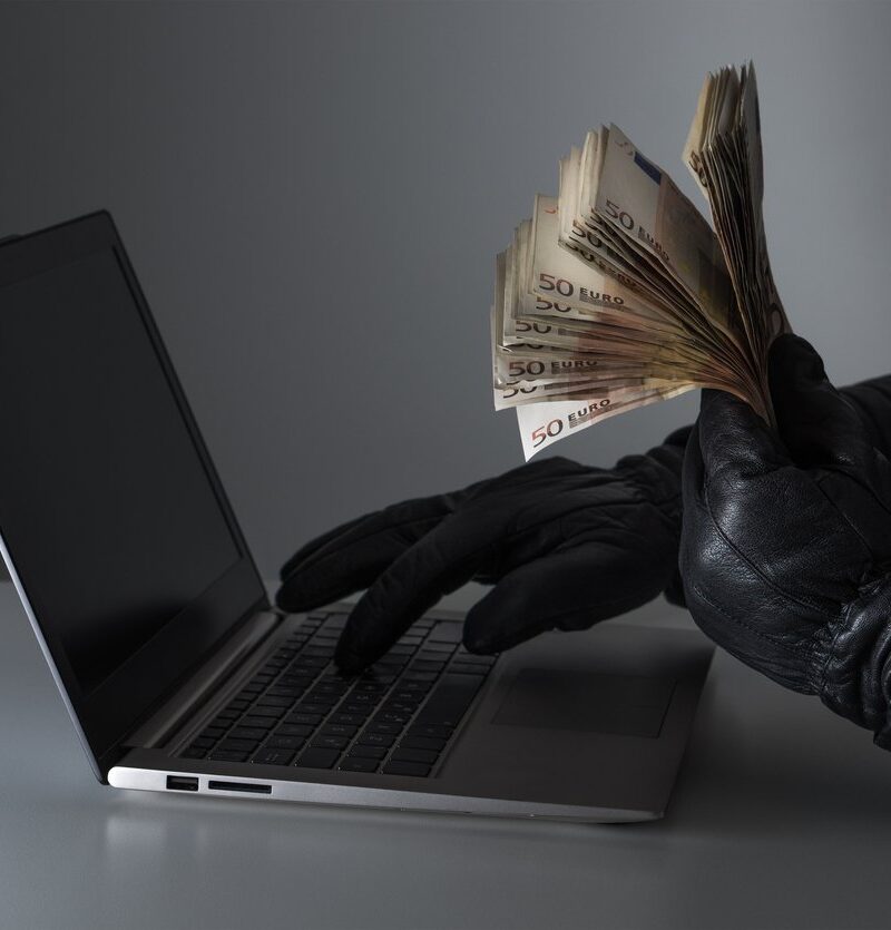 conceito de burla informatica com um criminoso com muito dinheiro na mão e a utilizar um computador portátil com luvas de cabedal pretas.