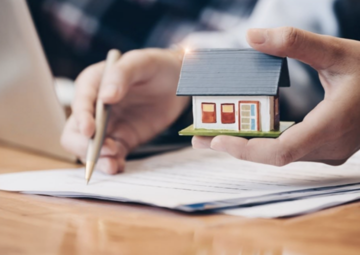 Escritura de imóvel com a casa numa mão e a caneta na outra mão.