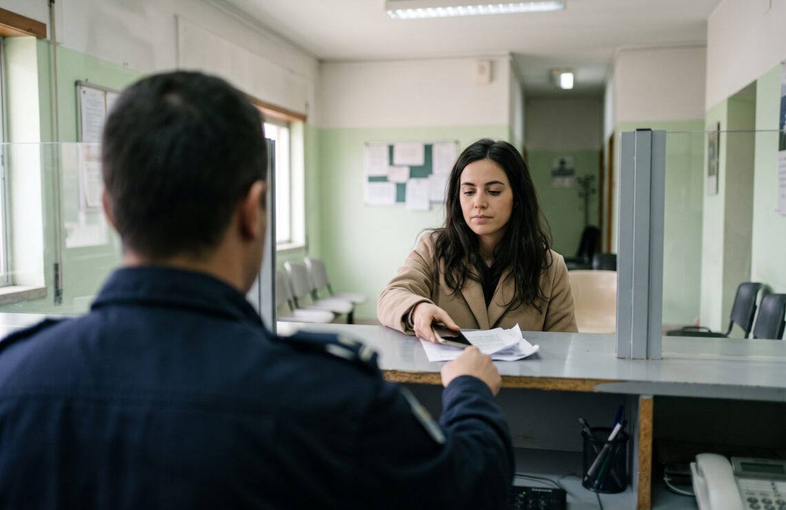 Mulher sentada a um balcão de atendimento numa esquadra em Portugal, a entregar um telemóvel e papéis a um agente de costas para a câmara