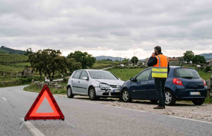 Dois carros imobilizados na berma de uma estrada após colisão, com triângulo de sinalização colocado e condutor ao telemóvel.