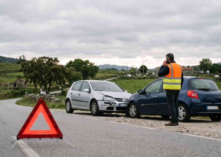 Dois carros imobilizados na berma de uma estrada após colisão, com triângulo de sinalização colocado e condutor ao telemóvel.
