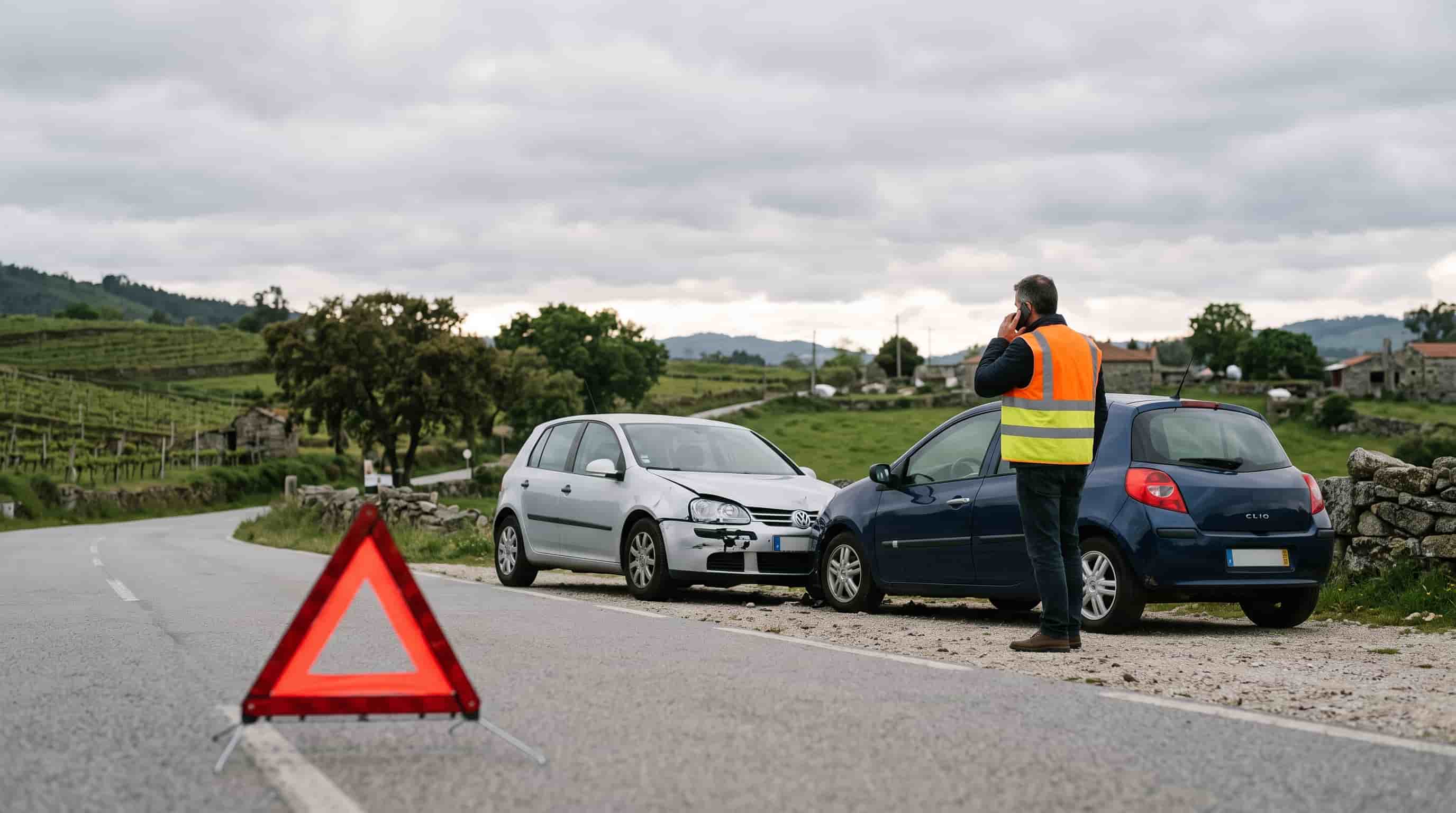 Dois carros imobilizados na berma de uma estrada após colisão, com triângulo de sinalização colocado e condutor ao telemóvel.