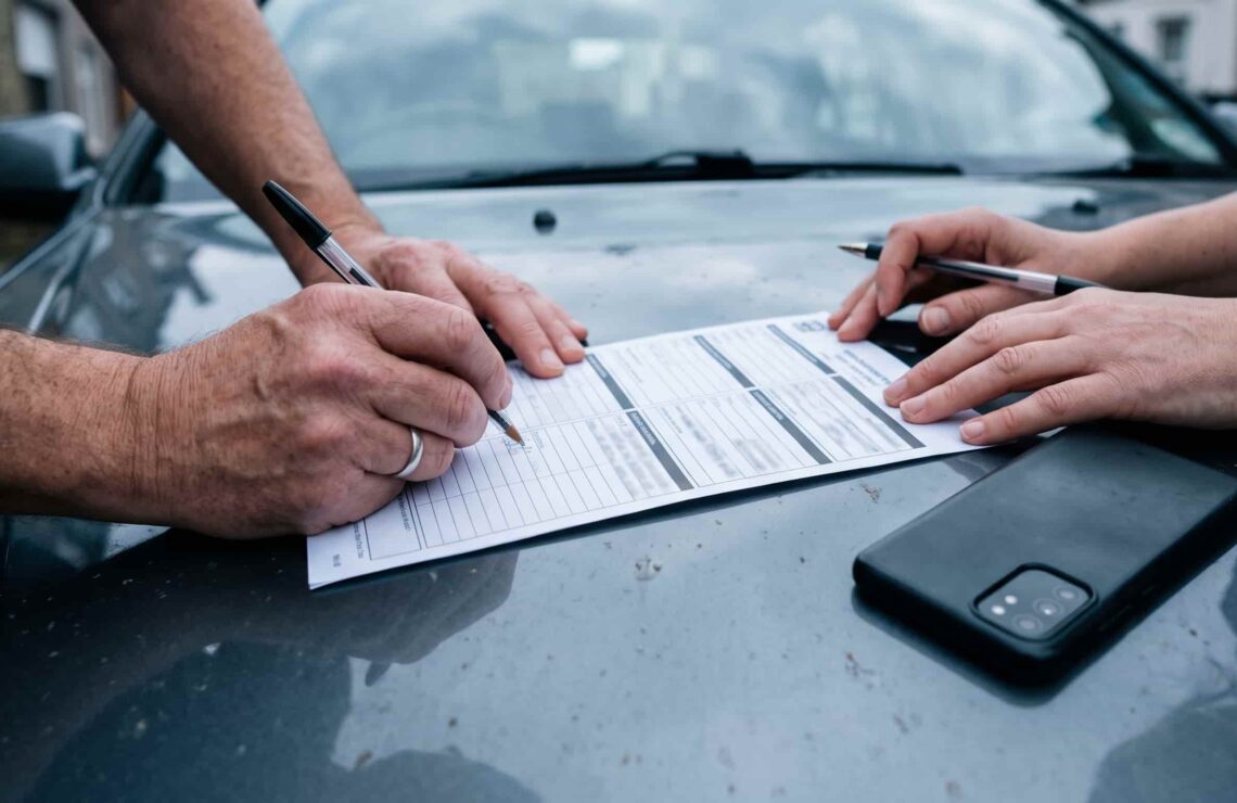Mãos de dois condutores a preencher a declaração amigável sobre o capô de um carro, com um telemóvel a fotografar os danos ao lado.