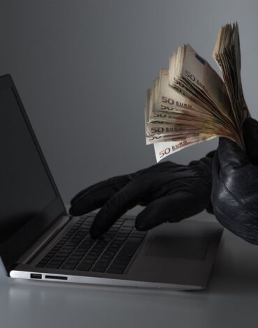 internet fraud and cyber security concept. criminal holding a lot of money and using laptop with black leather gloves.