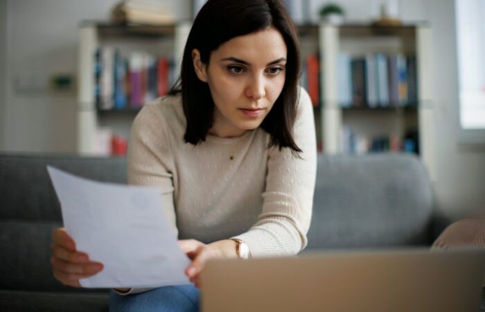 Mulher (senhoria empresária) com expressão preocupada sentada no sofá, a ler documentos e a olhar para o ecrã de um portátil, refletindo sobre o atraso da renda do inquilino