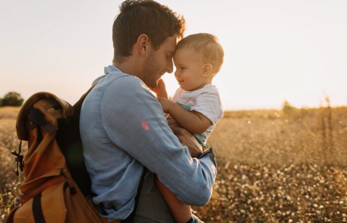 Pai e filho pequenos abraçados carinhosamente num campo dourado ao pôr do sol, a desfrutar de um momento de ligação e afeto. O pai carrega uma mochila e tem um sorriso suave enquanto o filho, ao colo, retribui o carinho. Evoca paternidade ativa e felicidade familiar.