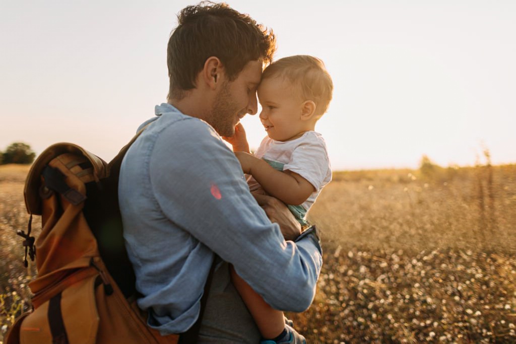 Pai e filho pequenos abraçados carinhosamente num campo dourado ao pôr do sol, a desfrutar de um momento de ligação e afeto. O pai carrega uma mochila e tem um sorriso suave enquanto o filho, ao colo, retribui o carinho. Evoca paternidade ativa e felicidade familiar.