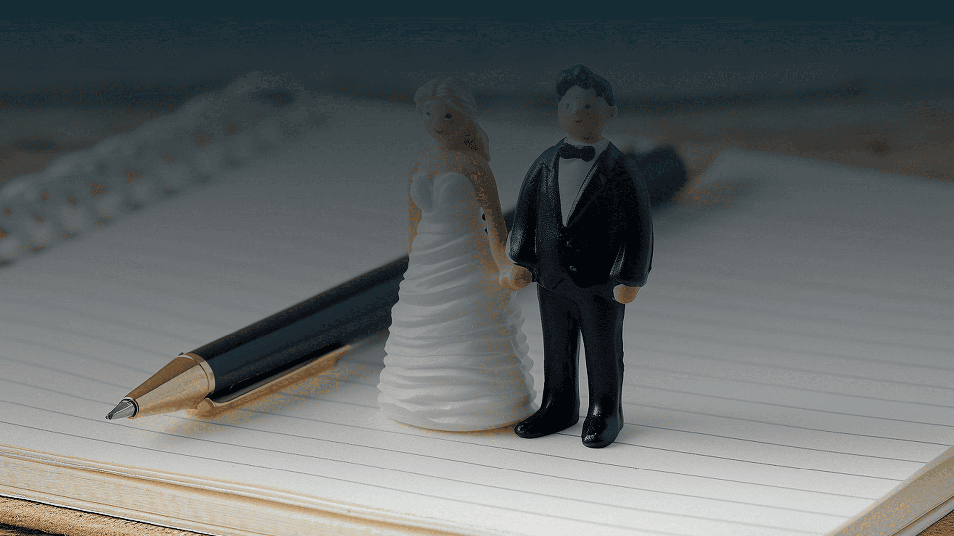 Pequenas figuras de um casal de noivos em cima de um caderno de notas aberto com uma caneta, simbolizando o planeamento legal do casamento.