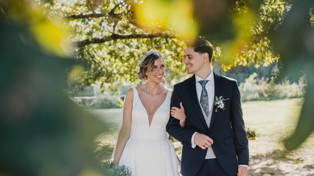 Noiva sorridente abraçada pelo noivo num ambiente de jardim, com folhagens verdes ao fundo.