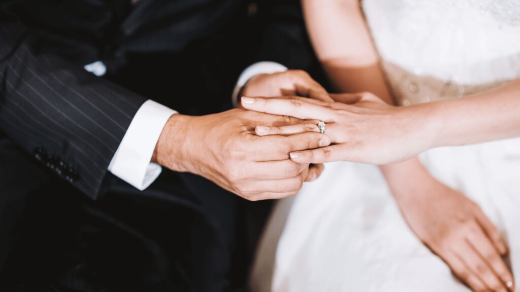 Pormenor das mãos de um casal durante a troca de alianças, com o noivo a colocar o anel no dedo da noiva.
