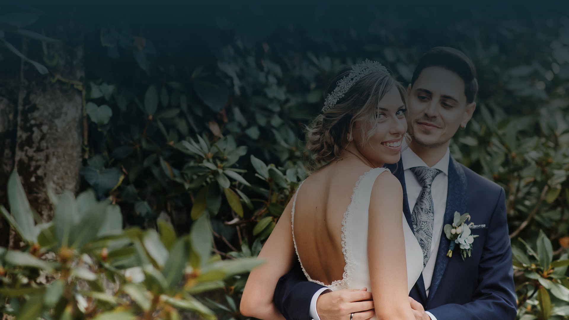 Noiva sorridente abraçada pelo noivo num ambiente de jardim, com folhagens verdes ao fundo.