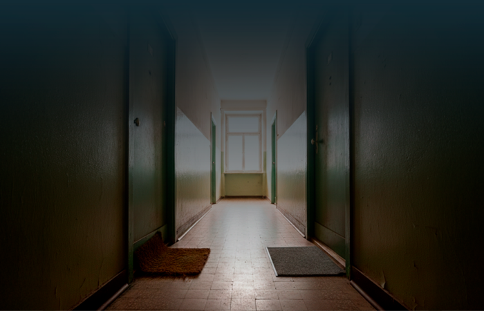 Corredor de prédio com duas portas fechadas lado a lado, cada uma com um tapete diferente à frente, iluminadas por luz natural ao fundo.