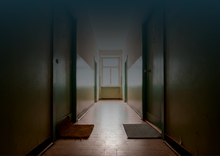 Corredor de prédio com duas portas fechadas lado a lado, cada uma com um tapete diferente à frente, iluminadas por luz natural ao fundo.