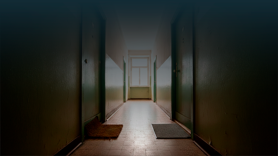 Corredor de prédio com duas portas fechadas lado a lado, cada uma com um tapete diferente à frente, iluminadas por luz natural ao fundo.