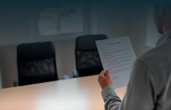 Pessoa vista de costas a segurar um contrato de trabalho com várias cláusulas sublinhadas a vermelho, simbolizando a análise de termos ilegais ou abusivos.