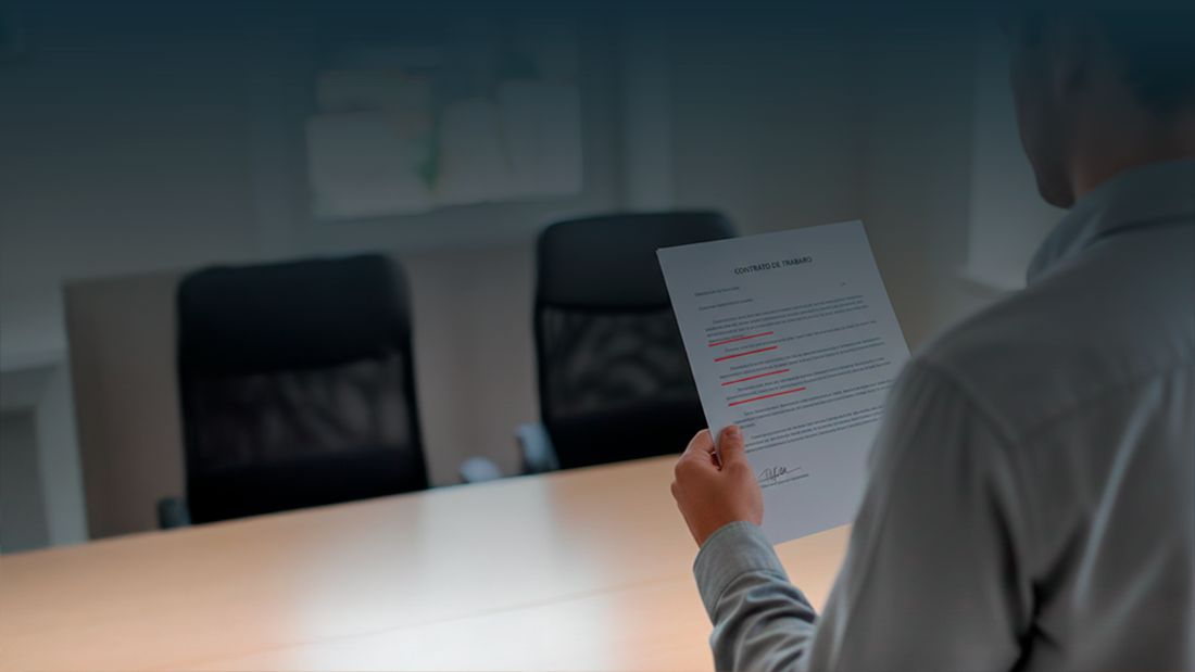Pessoa vista de costas a segurar um contrato de trabalho com várias cláusulas sublinhadas a vermelho, simbolizando a análise de termos ilegais ou abusivos.