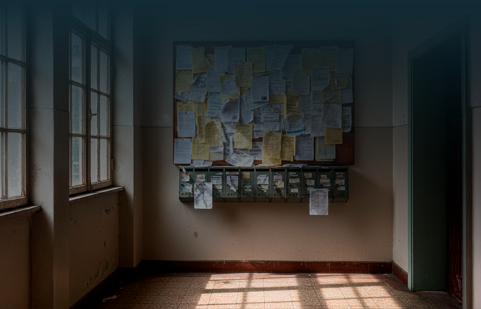 Hall de entrada de um prédio com caixas de correio antigas e um quadro de avisos cheio de papéis sobrepostos, comunicados riscados e notas desalinhadas.
