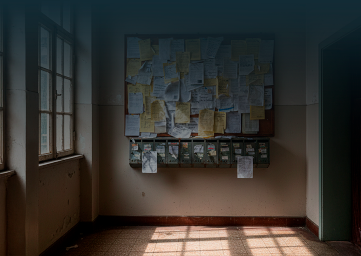 Hall de entrada de um prédio com caixas de correio antigas e um quadro de avisos cheio de papéis sobrepostos, comunicados riscados e notas desalinhadas.