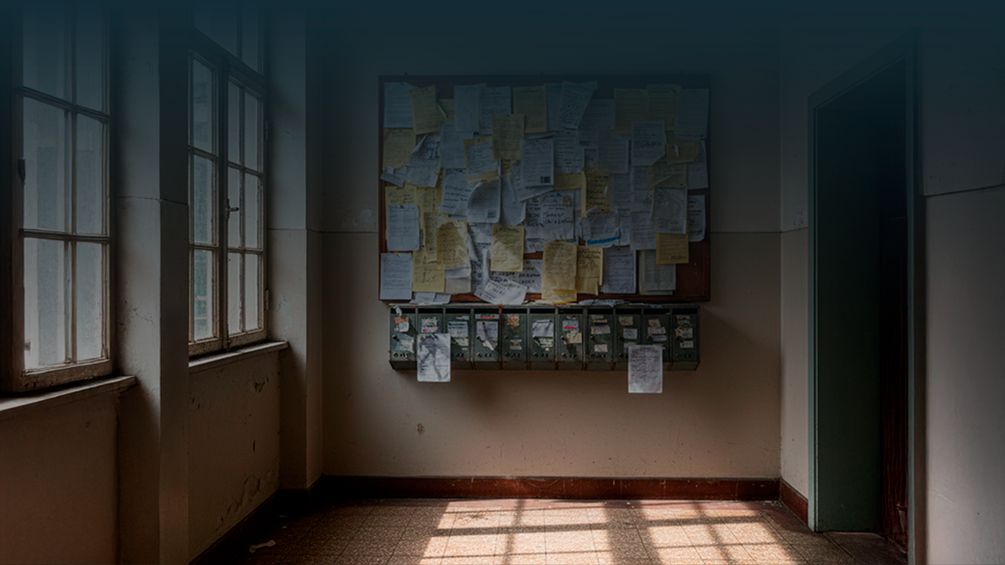Hall de entrada de um prédio com caixas de correio antigas e um quadro de avisos cheio de papéis sobrepostos, comunicados riscados e notas desalinhadas.