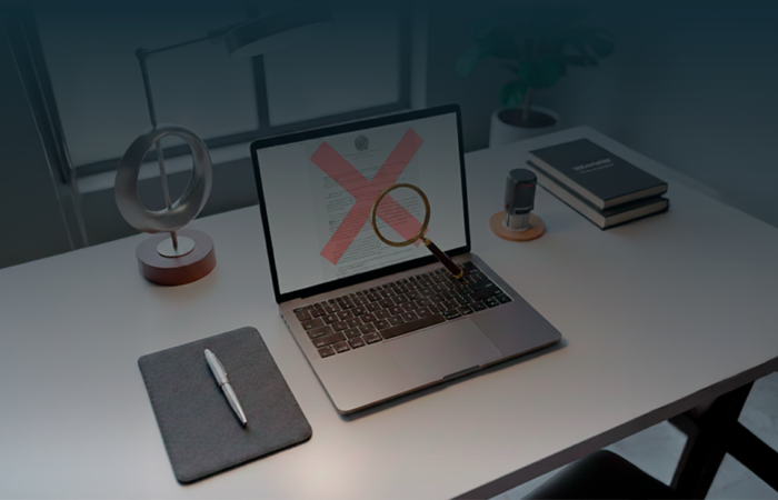 Portátil aberto exibindo edital de concurso público com um 'X' vermelho de erro e uma lupa sobre a mesa, simbolizando a análise de irregularidades em documentos oficiais.