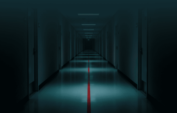 Um corredor longo e vazio, visto em perspetiva central, com portas numeradas de ambos os lados. No chão, uma única linha vermelha avança pelo corredor e desaparece ao longe. A iluminação é irregular: zonas de luz alternam com sombra, sugerindo avanço progressivo. Não há sinal de urgência imediata, apenas continuidade inevitável. Imagem de capa editorial para a pergunta: o que acontece quando nada é feito.