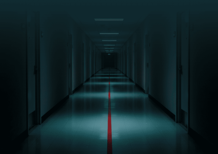 Um corredor longo e vazio, visto em perspetiva central, com portas numeradas de ambos os lados. No chão, uma única linha vermelha avança pelo corredor e desaparece ao longe. A iluminação é irregular: zonas de luz alternam com sombra, sugerindo avanço progressivo. Não há sinal de urgência imediata, apenas continuidade inevitável. Imagem de capa editorial para a pergunta: o que acontece quando nada é feito.