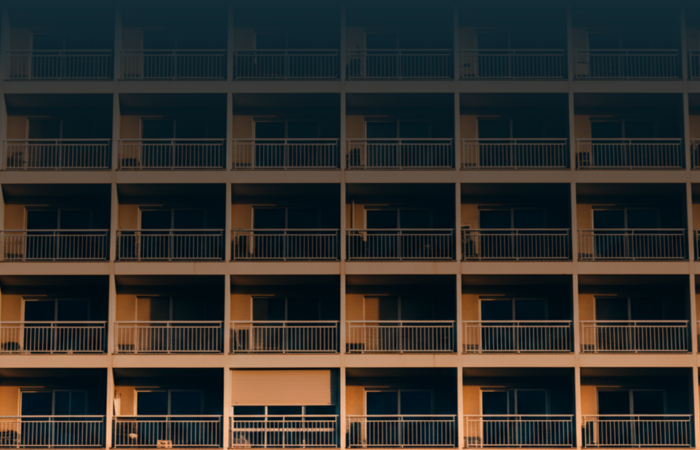 Fachada de prédio residencial vista de baixo, com várias varandas alinhadas e uma persiana parcialmente fechada, iluminada pela luz do final do dia.