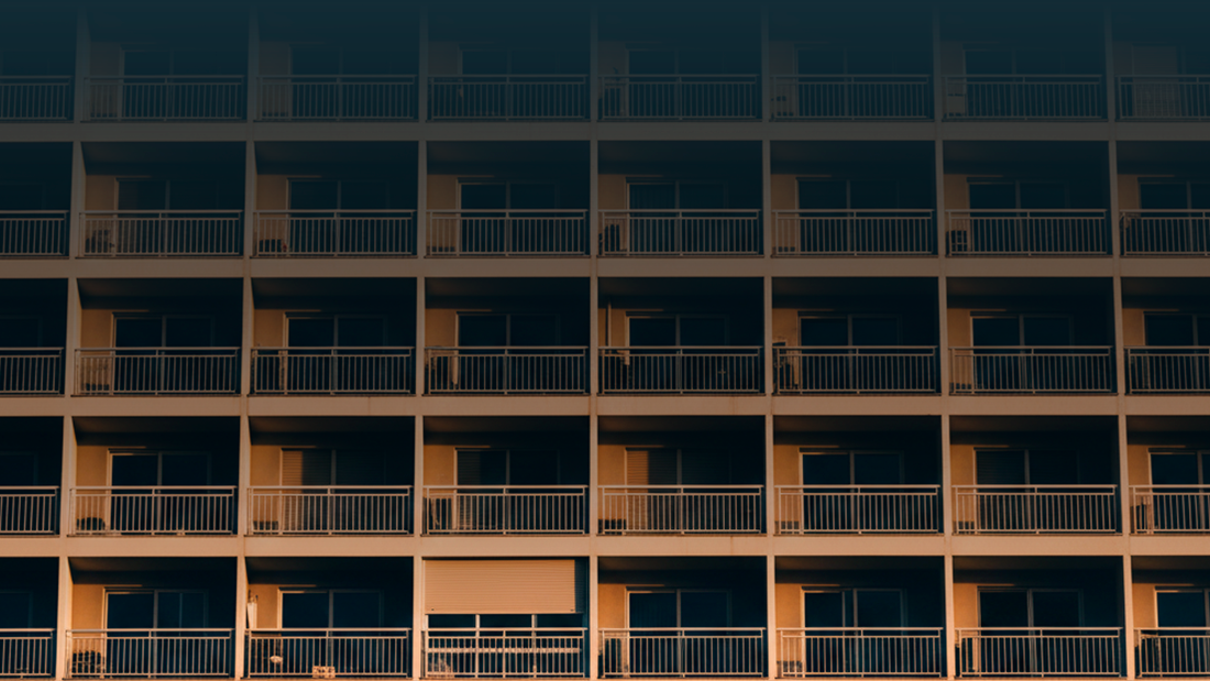 Fachada de prédio residencial vista de baixo, com várias varandas alinhadas e uma persiana parcialmente fechada, iluminada pela luz do final do dia.