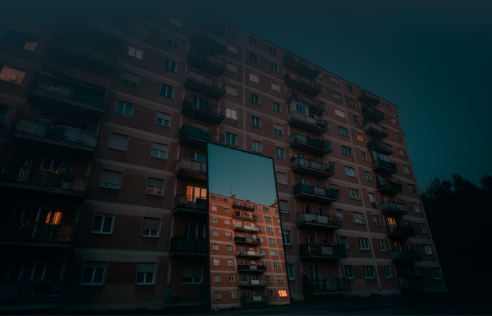 Edifício de habitação coletiva visto de baixo, com várias varandas iluminadas, refletido num espelho colocado ao nível do solo em frente à fachada.