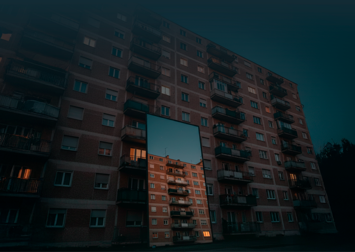 Edifício de habitação coletiva visto de baixo, com várias varandas iluminadas, refletido num espelho colocado ao nível do solo em frente à fachada.