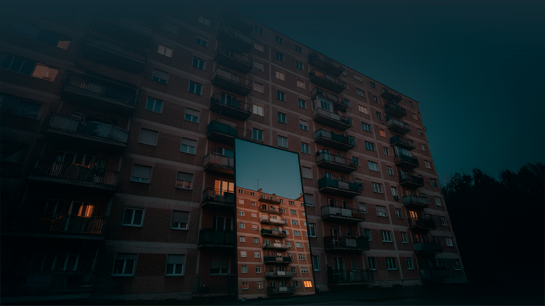 Edifício de habitação coletiva visto de baixo, com várias varandas iluminadas, refletido num espelho colocado ao nível do solo em frente à fachada.