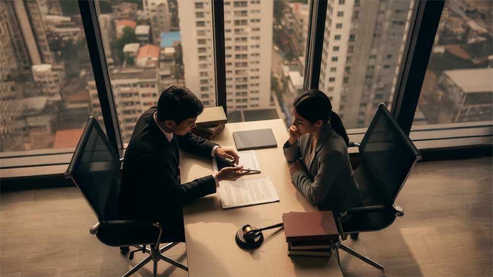 Vista aérea de uma reunião entre um advogado e uma cliente, com documentos e livros sobre a mesa, num escritório com grandes janelas e luz de entardecer.
