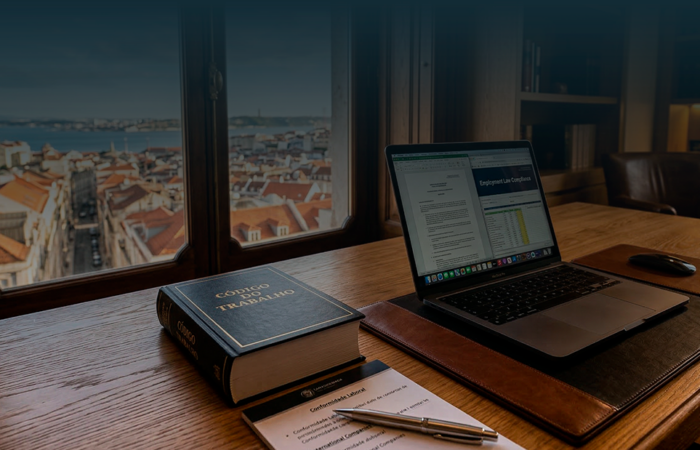 Close-up de uma secretária de carvalho com um livro do 'Código do Trabalho' português e um portátil aberto, em frente a uma janela com vista para os telhados de terracota de uma cidade em Portugal.