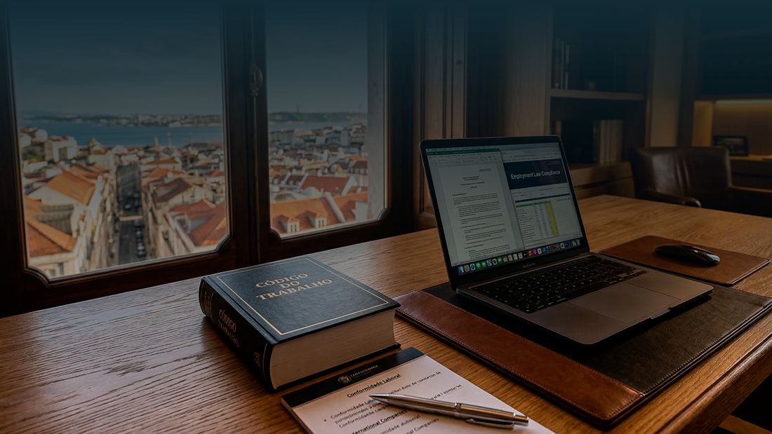 Close-up de uma secretária de carvalho com um livro do 'Código do Trabalho' português e um portátil aberto, em frente a uma janela com vista para os telhados de terracota de uma cidade em Portugal.