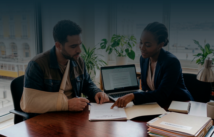 Um jovem trabalhador com o braço numa tipóia bege está sentado a uma mesa de escritório, a assinar documentos. Ao seu lado, uma advogada negra, com um blazer azul, aponta para o papel e explica o processo. Ao fundo, uma janela ampla revela uma vista urbana de Lisboa com um elétrico amarelo.