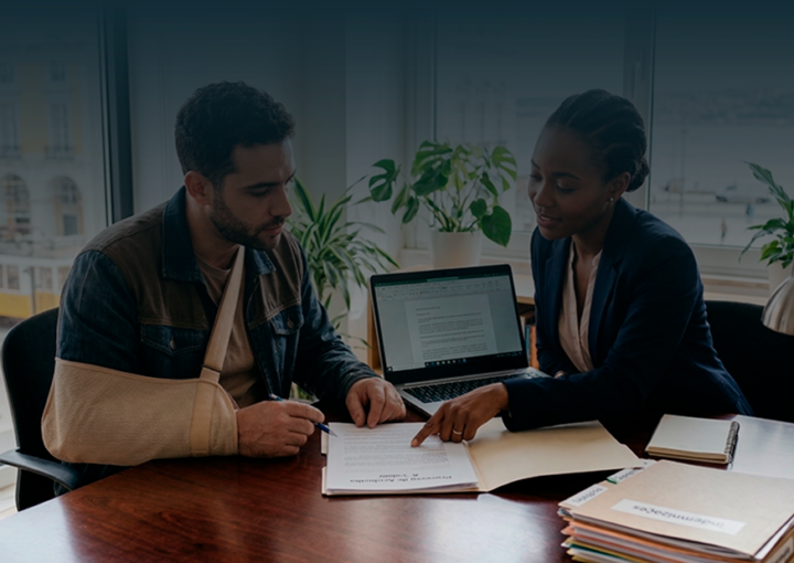 Um jovem trabalhador com o braço numa tipóia bege está sentado a uma mesa de escritório, a assinar documentos. Ao seu lado, uma advogada negra, com um blazer azul, aponta para o papel e explica o processo. Ao fundo, uma janela ampla revela uma vista urbana de Lisboa com um elétrico amarelo.