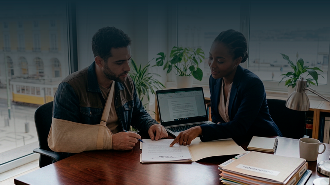 Um jovem trabalhador com o braço numa tipóia bege está sentado a uma mesa de escritório, a assinar documentos. Ao seu lado, uma advogada negra, com um blazer azul, aponta para o papel e explica o processo. Ao fundo, uma janela ampla revela uma vista urbana de Lisboa com um elétrico amarelo.