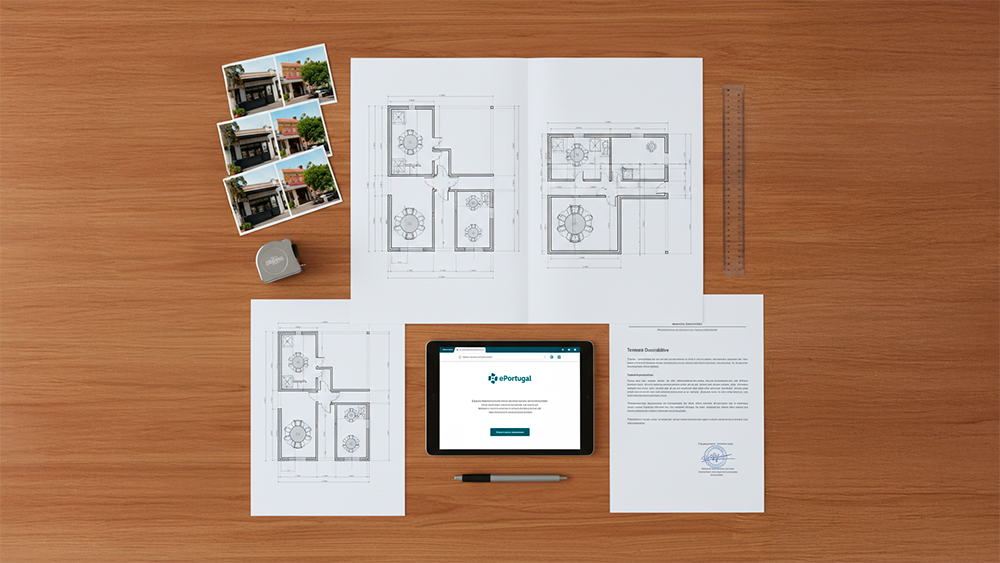 Vista superior de uma mesa de trabalho com plantas arquitetónicas de uma esplanada, memória descritiva, canetas e um tablet, simbolizando a fase de instrução de um processo de legalização municipal.