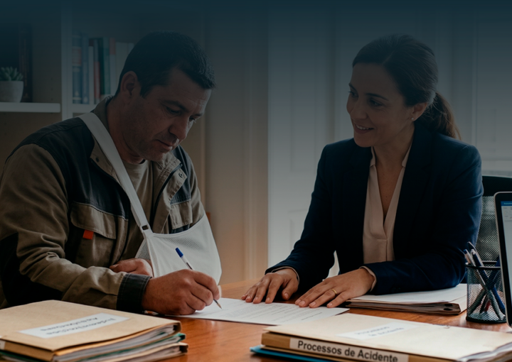 Uma advogada e o seu cliente, um homem com o braço numa tipóia, estão sentados numa mesa de escritório. Eles olham para documentos do processo de pedido de indemnização. Ambos têm expressões focadas e profissionais. O homem está a assinar um papel.