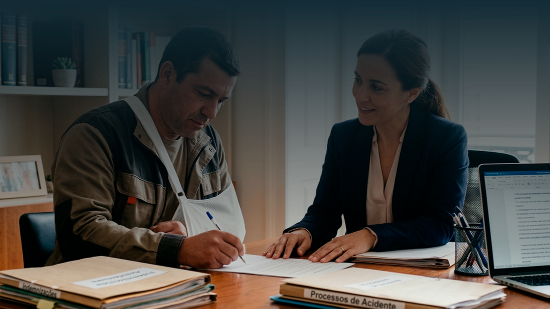 Uma advogada e o seu cliente, um homem com o braço numa tipóia, estão sentados numa mesa de escritório. Eles olham para documentos do processo de pedido de indemnização. Ambos têm expressões focadas e profissionais. O homem está a assinar um papel.