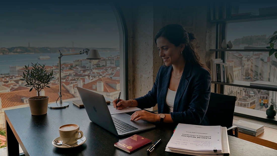 Empreendedora a trabalhar num escritório com vista para Lisboa, analisando documentos e portátil para abrir empresa em Portugal.
