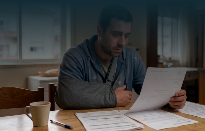 Homem com o braço ao peito numa tipoia, focado a ler um documento sobre indemnização por acidente de trabalho.
