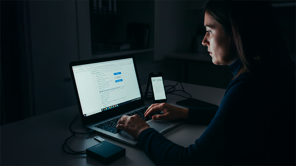 Fotografia de perfil de uma mulher num escritório iluminado pela luz do dia, sentada à secretária e focada no ecrã de um computador portátil. As suas mãos estão sobre o teclado, sugerindo a recolha e organização de ficheiros. Sobre a mesa, encontram-se um disco rígido externo e uma pen drive ligados ao computador, além de um smartphone num suporte exibindo um cronómetro. Ao fundo, vê-se uma estante com livros, criando um ambiente profissional de análise exaustiva e gestão de tempo.