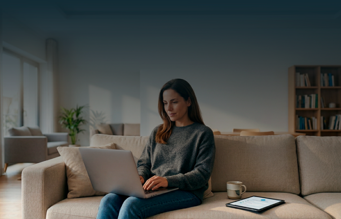 Uma mulher sentada de frente num sofá bege, a utilizar um portátil sobre o colo, num ambiente de sala de estar luminoso com estantes de livros ao fundo e texto sobreposto no topo.