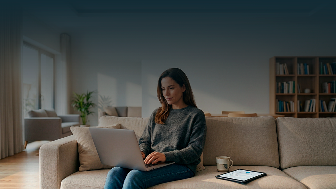 Uma mulher sentada de frente num sofá bege, a utilizar um portátil sobre o colo, num ambiente de sala de estar luminoso com estantes de livros ao fundo e texto sobreposto no topo.