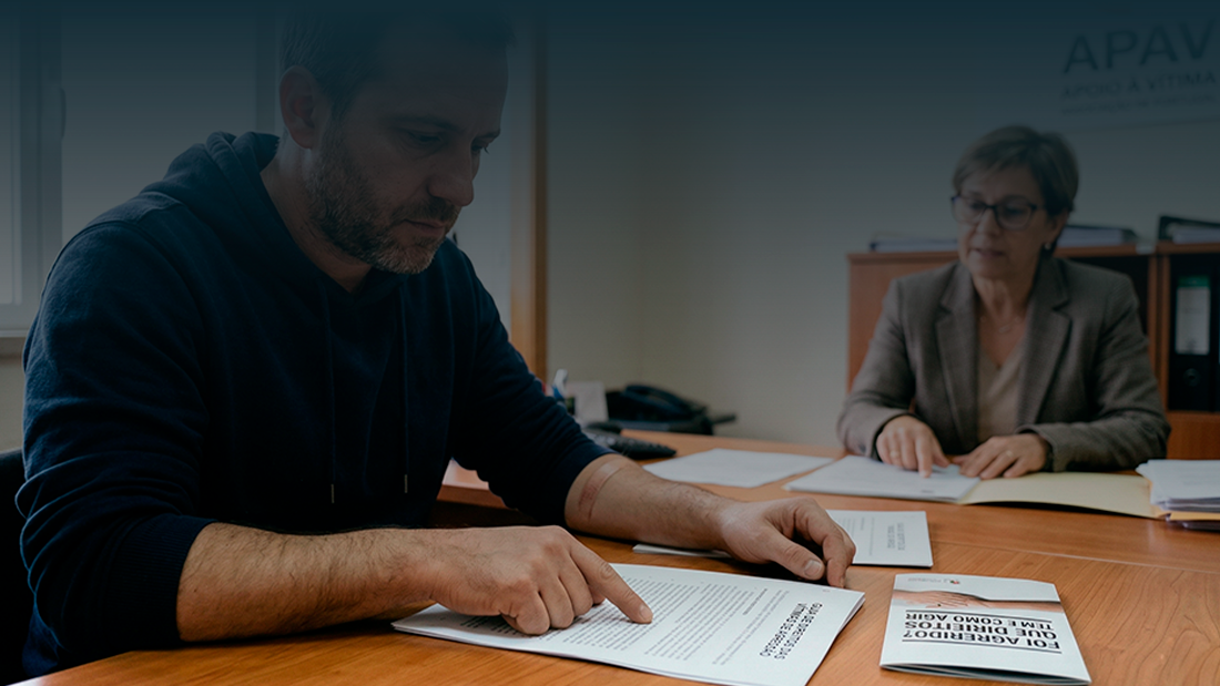 Um homem sentado num gabinete de apoio jurídico, focado na leitura de documentos sobre direitos das vítimas de agressão, com uma técnica especializada ao fundo num ambiente acolhedor e profissional.