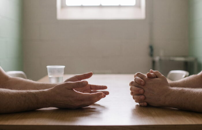 Duas pessoas sentadas frente a frente a uma mesa, com as mãos pousadas no tampo, numa sala de paredes claras.