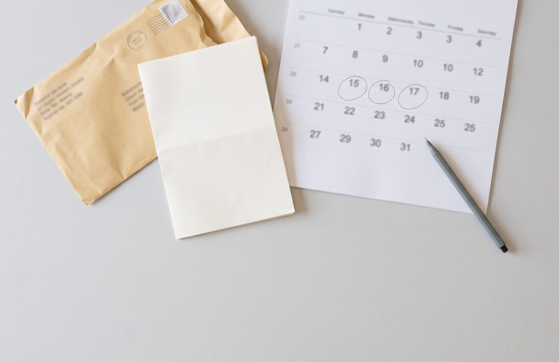 Envelope aberto, folha dobrada e calendário de parede pousados numa secretária, vistos de cima.