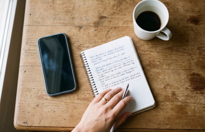 Mesa de cozinha em Portugal, de manhã, com um telemóvel, um caderno aberto com anotações manuscritas e uma caneca de café, vista de cima