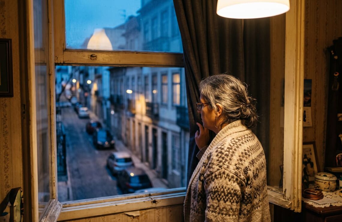 Mulher de meia-idade a olhar pela janela de um apartamento para uma rua residencial portuguesa, de pé, ao início da noite